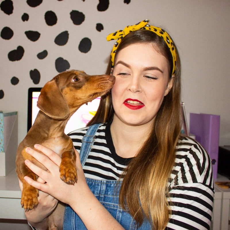 Holly from Sprinkle Club in a playful moment with her adorable dachshund, both set against a charming backdrop of black heart-shaped spots