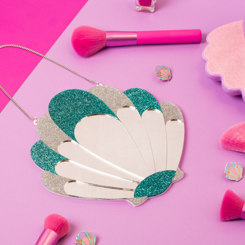 Glittery green and silver scallop shell-shaped wall mirror on a pink background with makeup accessories nearby