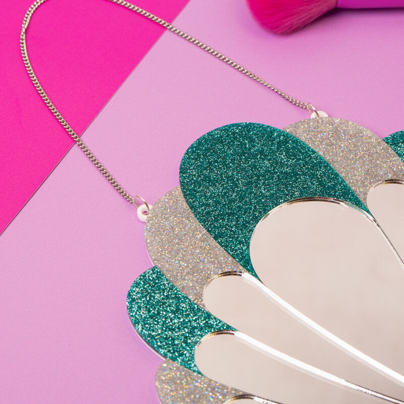 Close-up of a glittery scallop shell-shaped wall mirror with a chain for hanging, showcasing sparkling green and silver hues on a duo-tone pink background