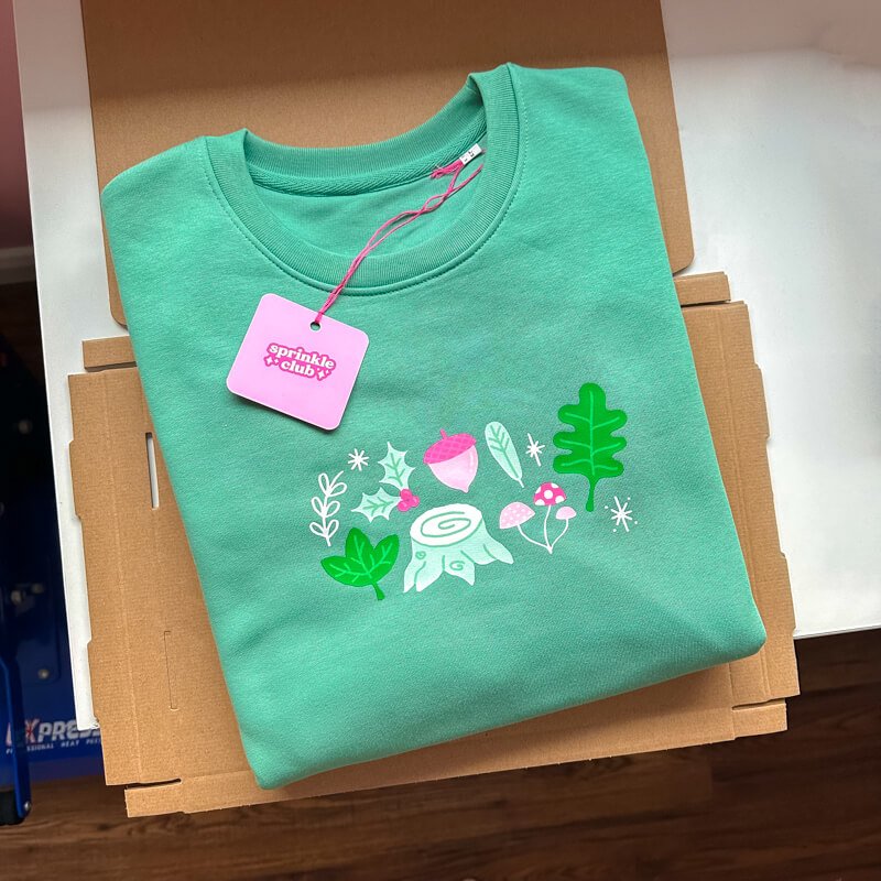 Sprinkle Club - A folded forest green sweatshirt with woodland items illustrated on the chest in pink and green