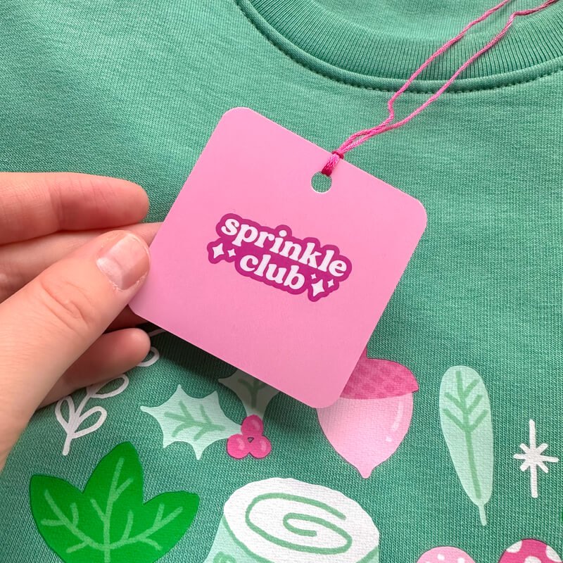 Sprinkle Club - A pink swing tag attached to a forest green sweatshirt with a cute woodland design