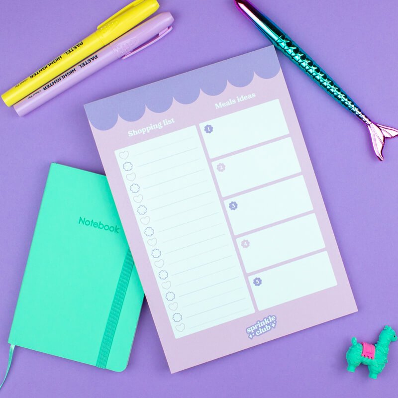 Pastel shopping list notepad with magnetic back, yellow and pink highlighters, blue mermaid tail pen, and green notebook on a purple background