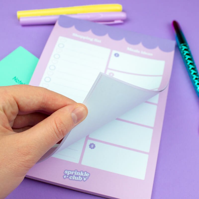 A hand peeling away a page on a pastel lilac magnetic shopping list notepad with scalloped edge design, placed on a purple background with colourful stationery items nearby