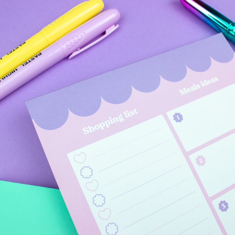 Pastel lilac magnetic shopping list notepad with scalloped edges, alongside yellow and teal stationery on a purple background