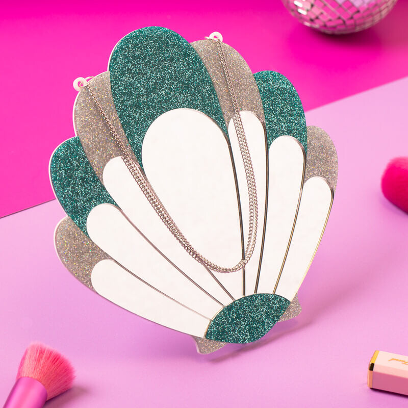 A vibrant and glittery scallop shell-shaped wall mirror with emerald and silver accents on a pink background, surrounded by makeup brushes