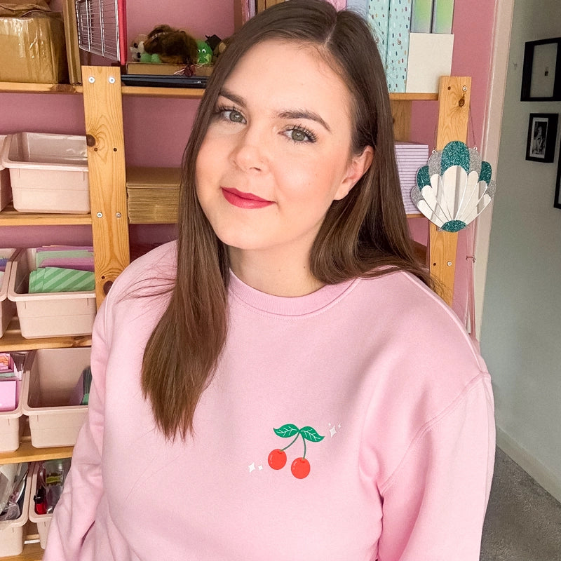 Holly from Sprinkle Club sporting a soft pink sweatshirt with a cute cherry design, complemented by her office's pastel decor and organised shelves in the background