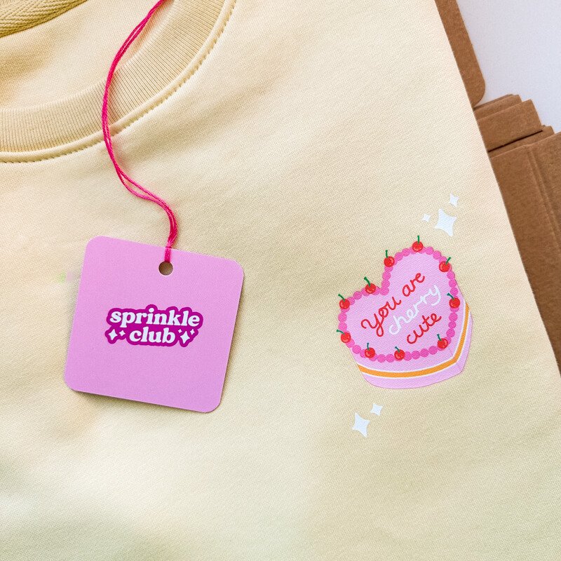 Sprinkle Club - A pastel yellow sweatshirt with a vintage cake illustration and a pink clothing swing tag
