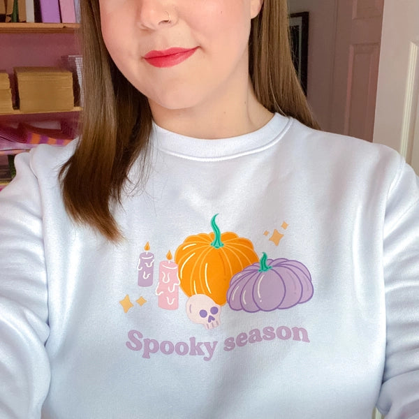 Sprinkle Club - A woman wearing a white illustrated halloween sweatshirt of a pumpkin and skull that says 'spooky season' in purple typography underneath