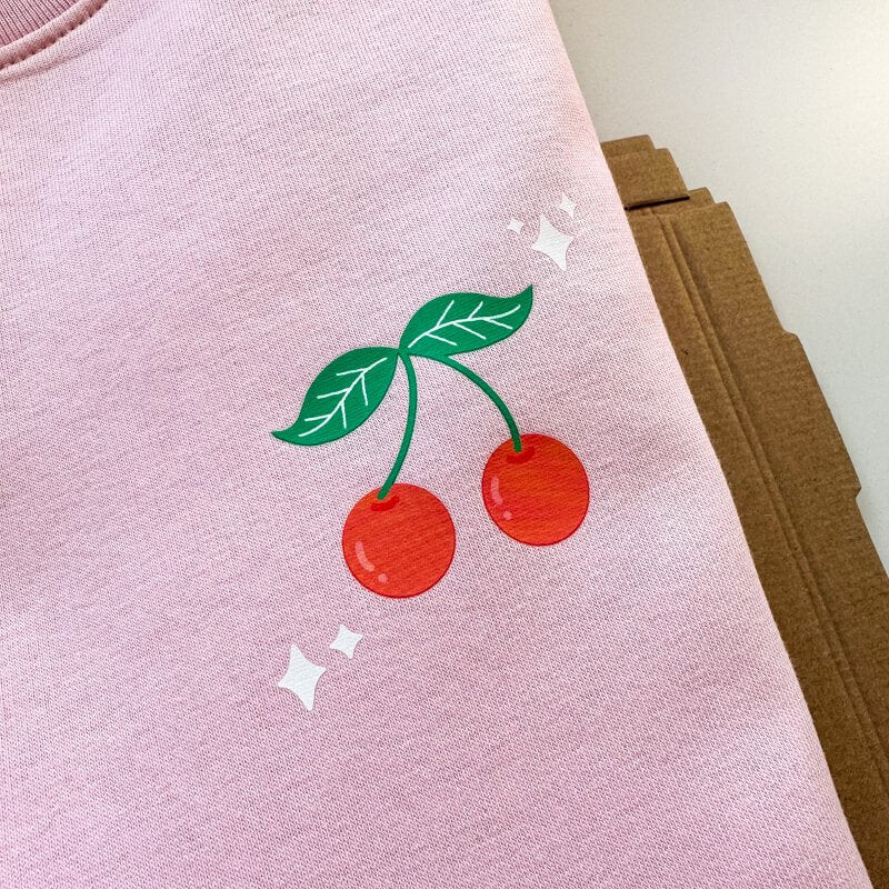 Sprinkle Club - Close up of a cute red cherry illustration on a pink sweatshirt