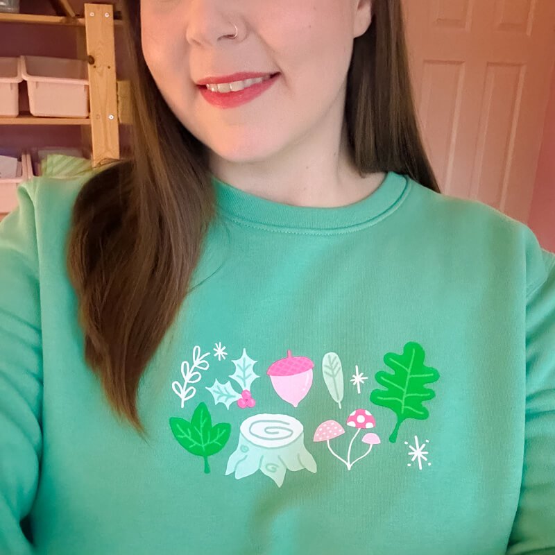 Sprinkle Club - A woman wearing a forest green sweatshirt with a cute illustration of forest items