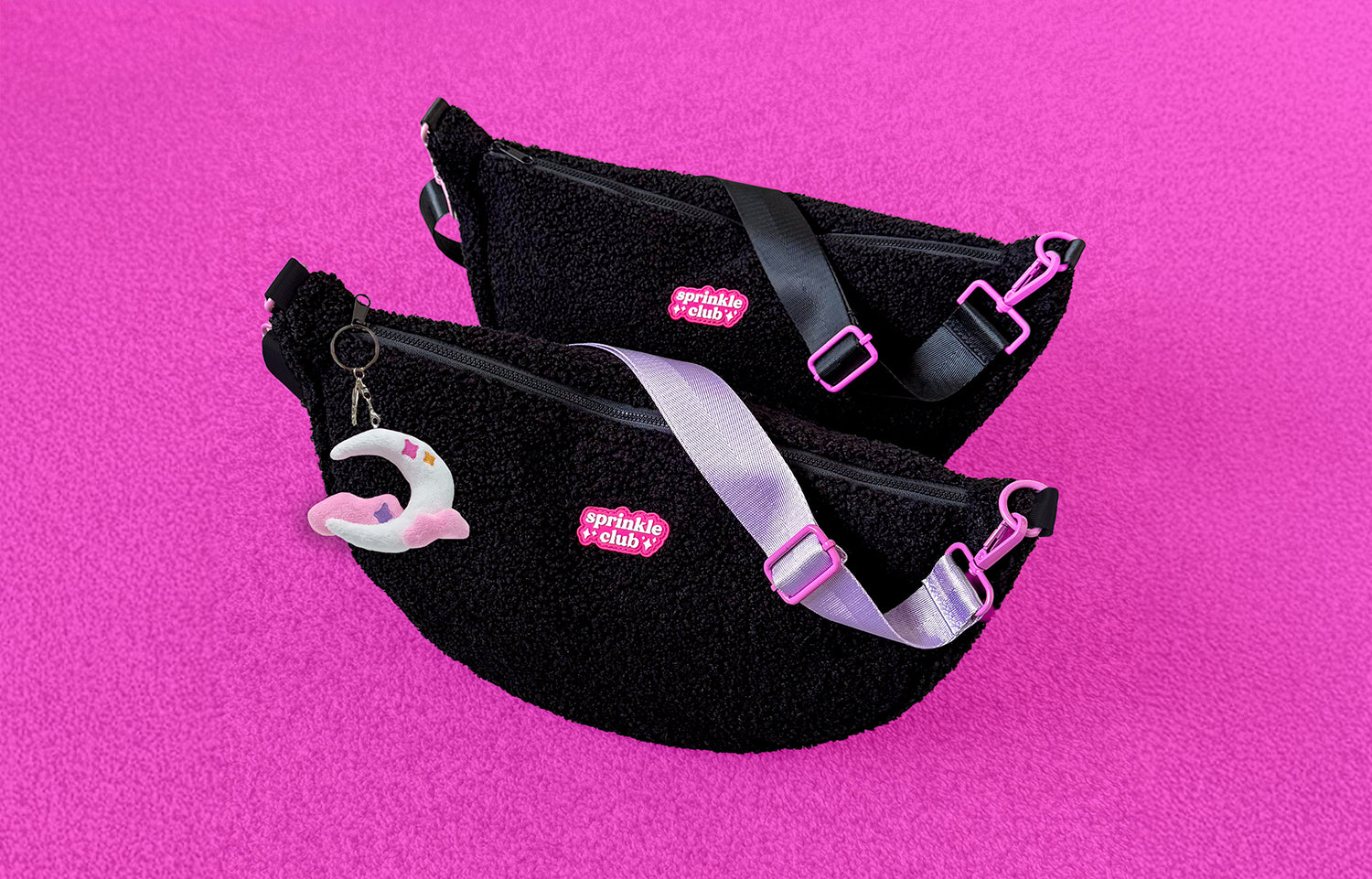 Sprinkle Club black sherpa sling bags with pink logo, shown with lilac and black straps and moon keychain, displayed on bright pink fabric