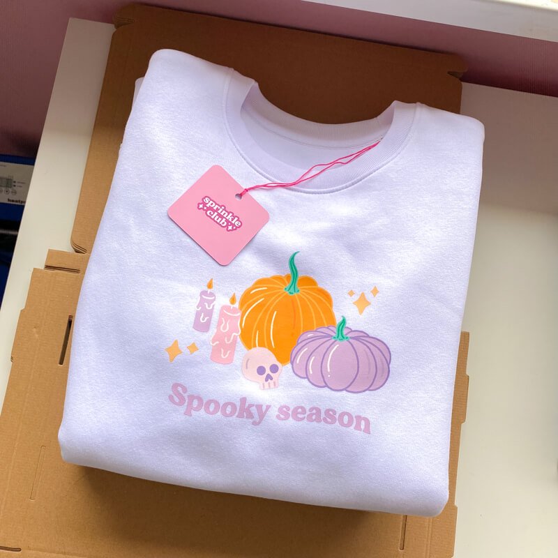 Sprinkle Club - A folded white halloween inspired sweatshirt that has a cute pumpkin and skull illustration on the chest