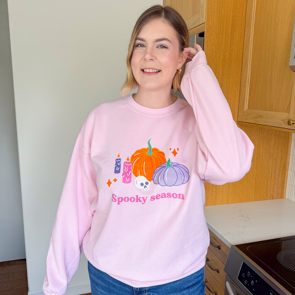 Woman smiling in the baby pink pastel Halloween pumpkin sweatshirt with bright pink Spooky season lettering, worn loose over jeans