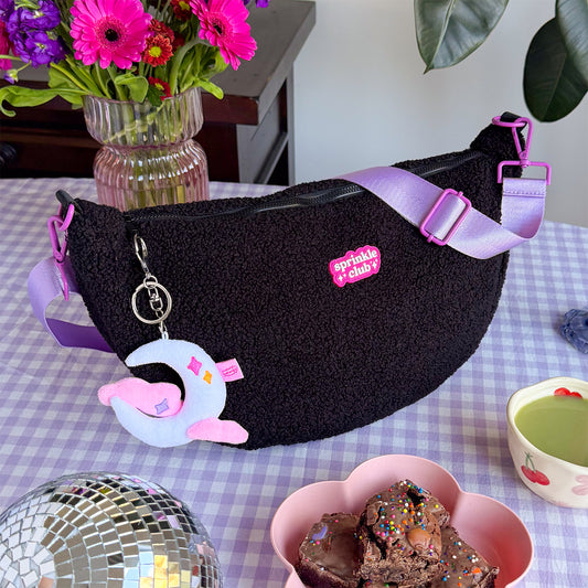 Black fluffy teddy crossbody bag with a lilac strap and moon plushie keychain, sitting on a lilac gingham tablecloth beside pink flowers, a disco ball and a bowl of sprinkle-topped brownies