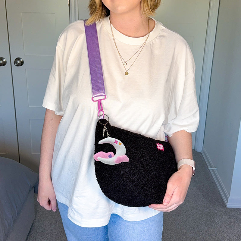 Woman wearing the black fluffy teddy crossbody bag across her body on a lilac strap, with the moon plushie keychain clipped on, styled with a white oversized t-shirt and jeans
