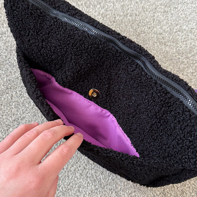 Hand opening the magnetic back pocket of a black fluffy teddy crossbody bag, showing the purple lining and gold-tone magnetic snap