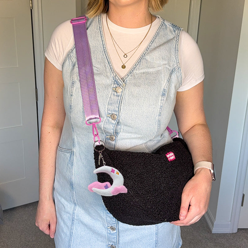 Woman wearing the black fluffy teddy crossbody bag on a lilac strap with the moon plushie keychain, styled with a denim pinafore dress over a white t-shirt