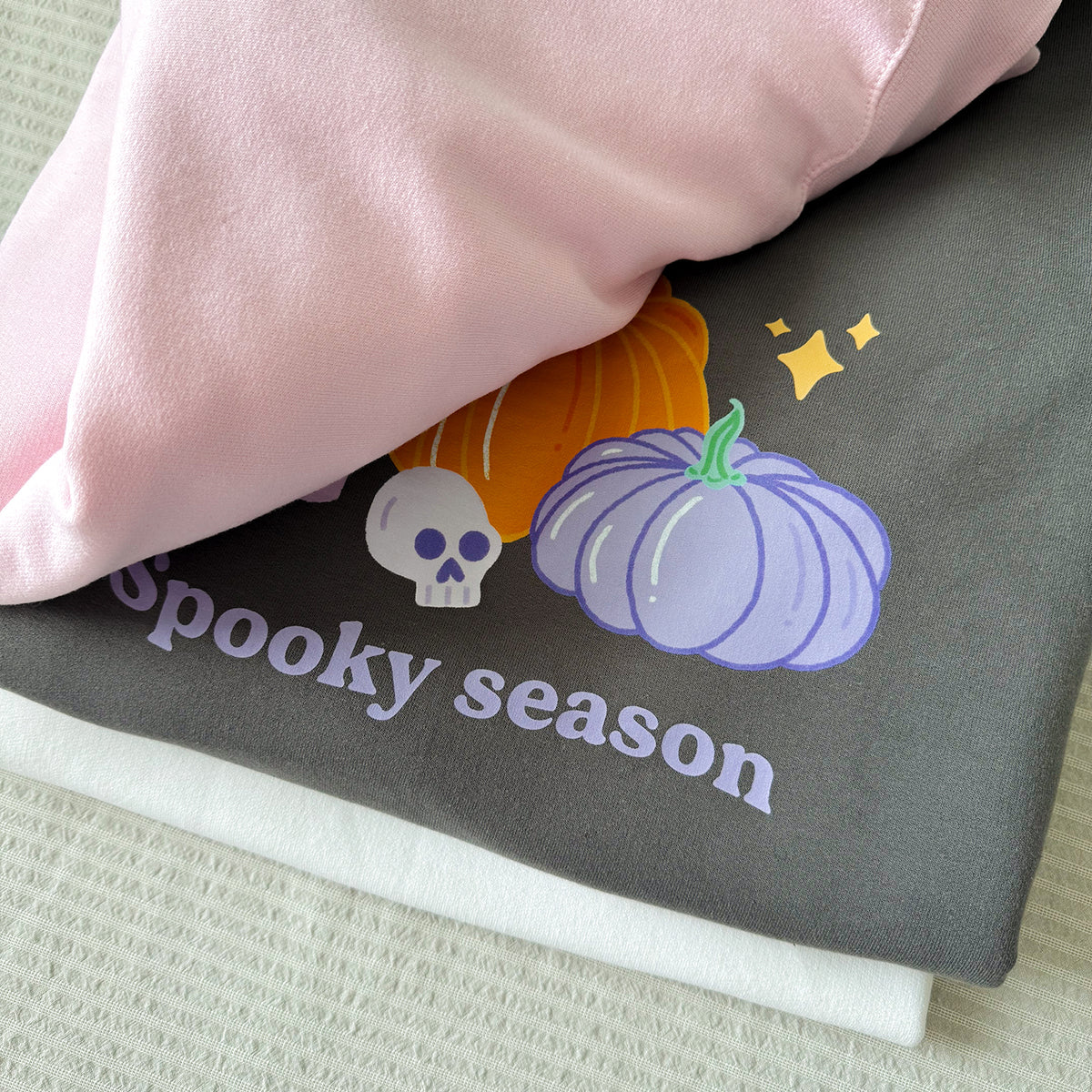 Close-up of the lilac Spooky season lettering, skull and pumpkins on the charcoal sweatshirt, with the baby pink and white colours folded alongside