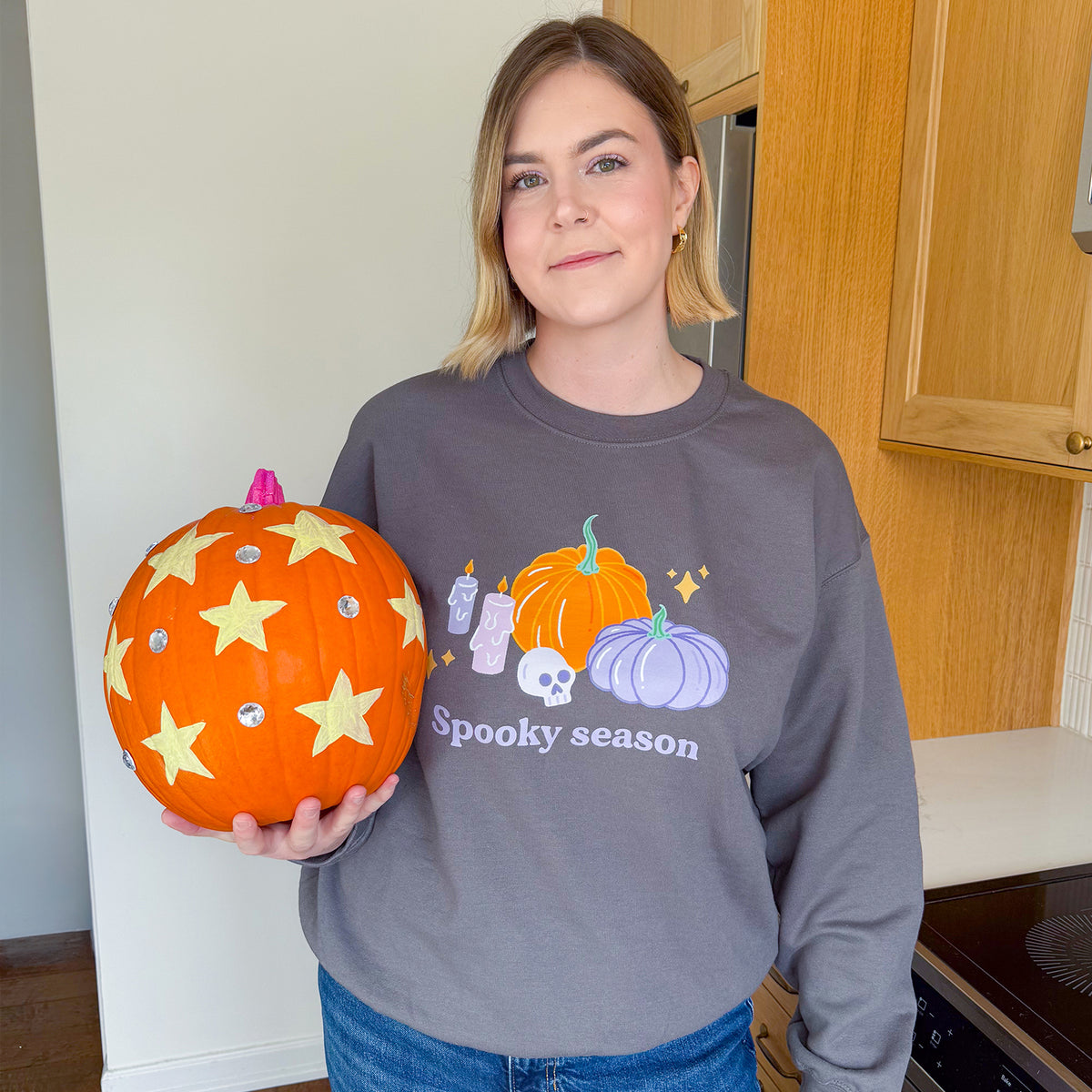 Woman wearing the charcoal pastel Halloween pumpkin sweatshirt with jeans, holding a real pumpkin painted with yellow stars and gems