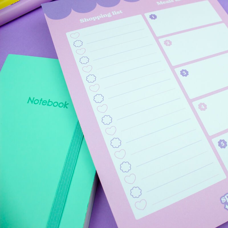 Pastel lilac shopping list notepad with adorable scalloped header and checklist, next to a mint green notebook on a purple surface