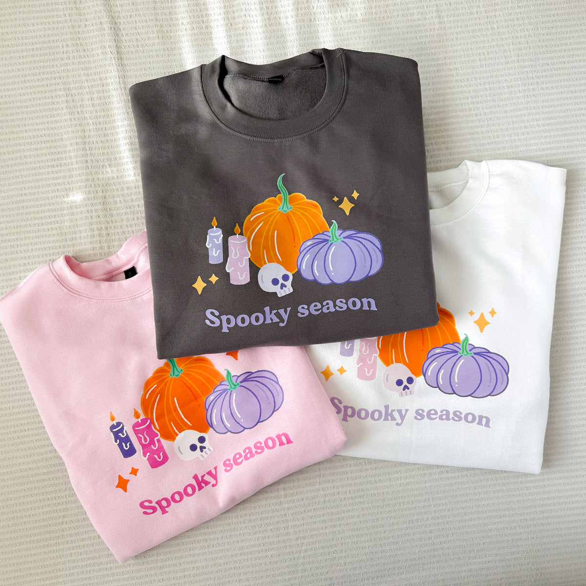 Three folded pastel Halloween pumpkin sweatshirts in baby pink, black and white, each printed with pumpkins, candles, a skull and Spooky season lettering