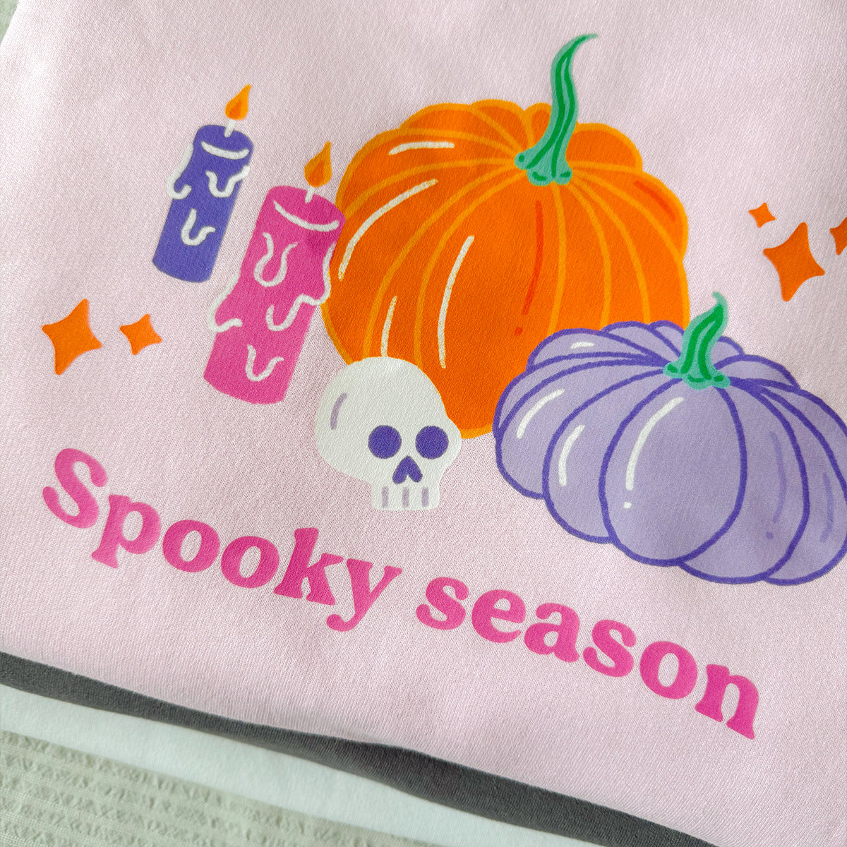 Close-up of the Spooky season print on the baby pink sweatshirt, showing orange and lilac pumpkins, purple and pink dripping candles and a white skull