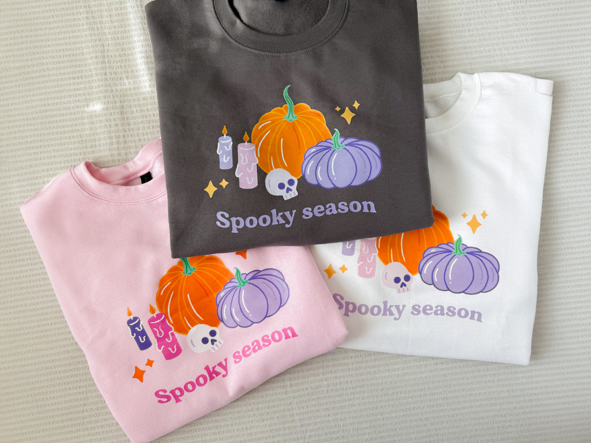 Pastel Halloween pumpkin sweatshirts in charcoal, baby pink and white laid out together, each printed with pumpkins, candles, a skull and Spooky season lettering