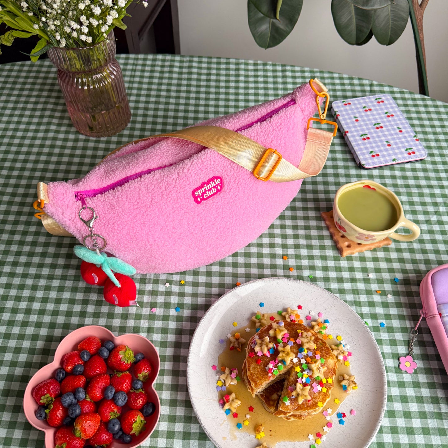 Top view of pink teddy crossbody bag styled on a green gingham table with pancakes, berries, Kindle and accessories