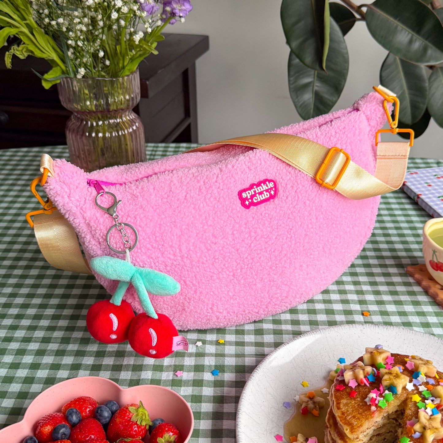 Pink teddy crossbody sling bag with cherry plushie keychain on a green gingham table