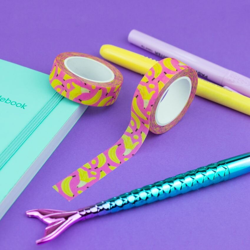 Stationery arrangement with pink and yellow banana fruit washi tape, a teal notebook, and colorful pens on a purple background