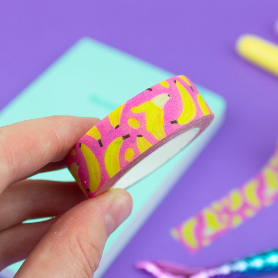 Close-up of a hand holding a roll of pink and yellow banana fruit washi tape against a purple background