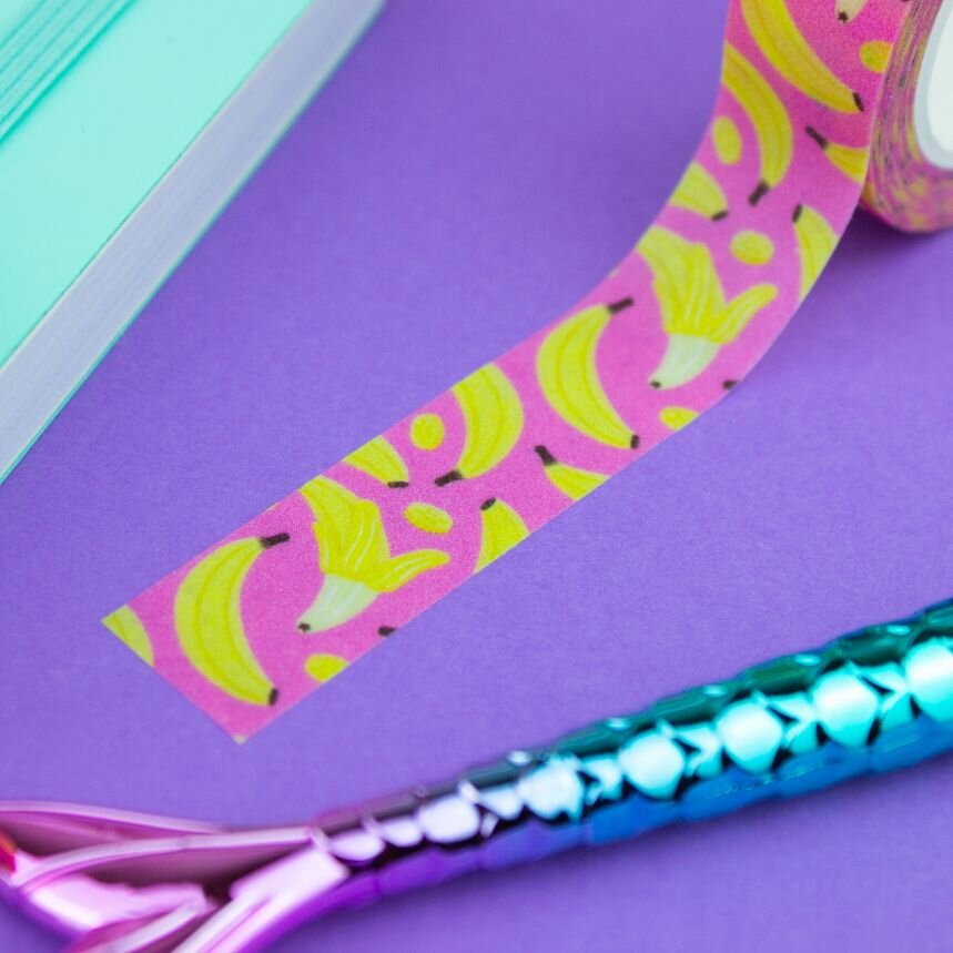 Diagonal strip of pink and yellow banana fruit washi tape alongside a teal mermaid tail pen on a purple background